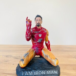 Pu&ograve; includere: Una statuetta di Iron Man rossa e dorata inginocchiata su una base nera con le parole "I AM IRON MAN" incise su di essa. La statuetta indossa una tuta danneggiata con crepe e graffi visibili. La statuetta alza la mano destra con il Guanto dell'Infinito.