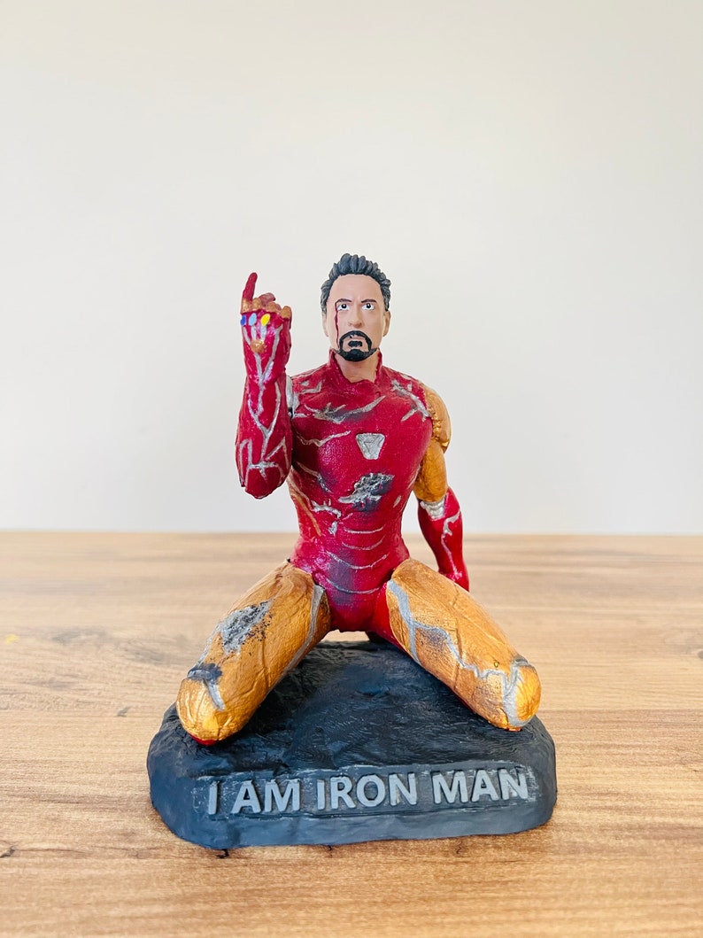 Pu&ograve; includere: Una statuetta di Iron Man rossa e dorata inginocchiata su una base nera con le parole "I AM IRON MAN" incise su di essa. La statuetta indossa una tuta danneggiata con crepe e graffi visibili. La statuetta alza la mano destra con il Guanto dell'Infinito.