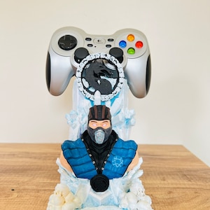 May include: A 3D printed figurine of Sub-Zero from Mortal Kombat, holding a video game controller. The figurine is set on a base that resembles ice and features a dragon symbol.