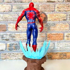 May include: A red and blue action figure of Spider-Man standing on a blue and brown base. The figure is posed with its back to the viewer, and its arms are outstretched.