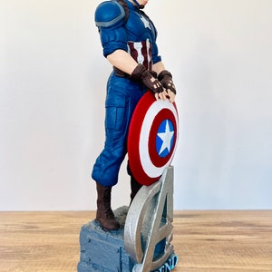 May include: A Captain America figurine in a dynamic pose, holding his iconic shield. The figure is clad in a blue and white costume, with a red, white, and blue shield. The base features the Avengers logo and the word "LEGEND".