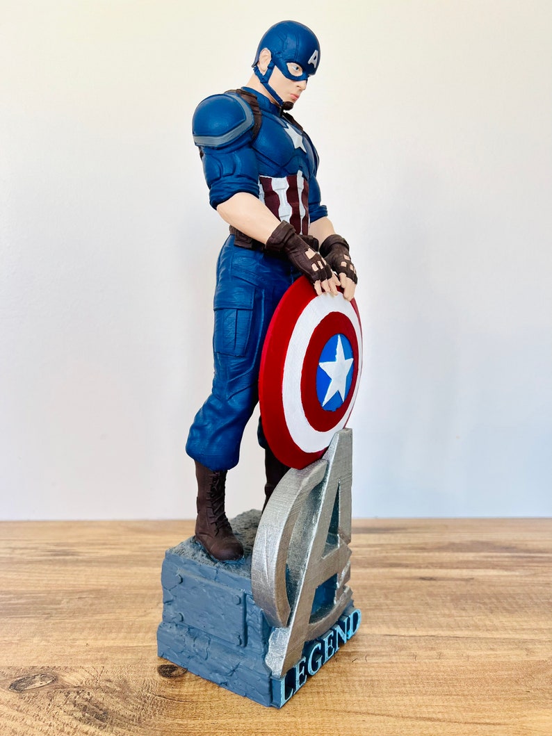 May include: A Captain America figurine in a dynamic pose, holding his iconic shield. The figure is clad in a blue and white costume, with a red, white, and blue shield. The base features the Avengers logo and the word "LEGEND".