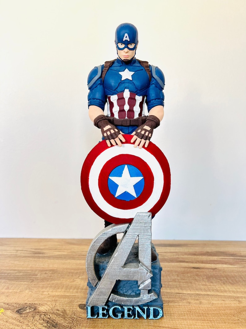 May include: A Captain America figurine in a blue, red, and white costume, holding a shield with a white star. The figurine stands on a base with the Avengers logo and the word "LEGEND" in blue.