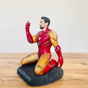 Pu&ograve; includere: Una action figure di Iron Man rossa e dorata inginocchiata su una base nera con le parole "I AM IRON MAN" incise sulla base.