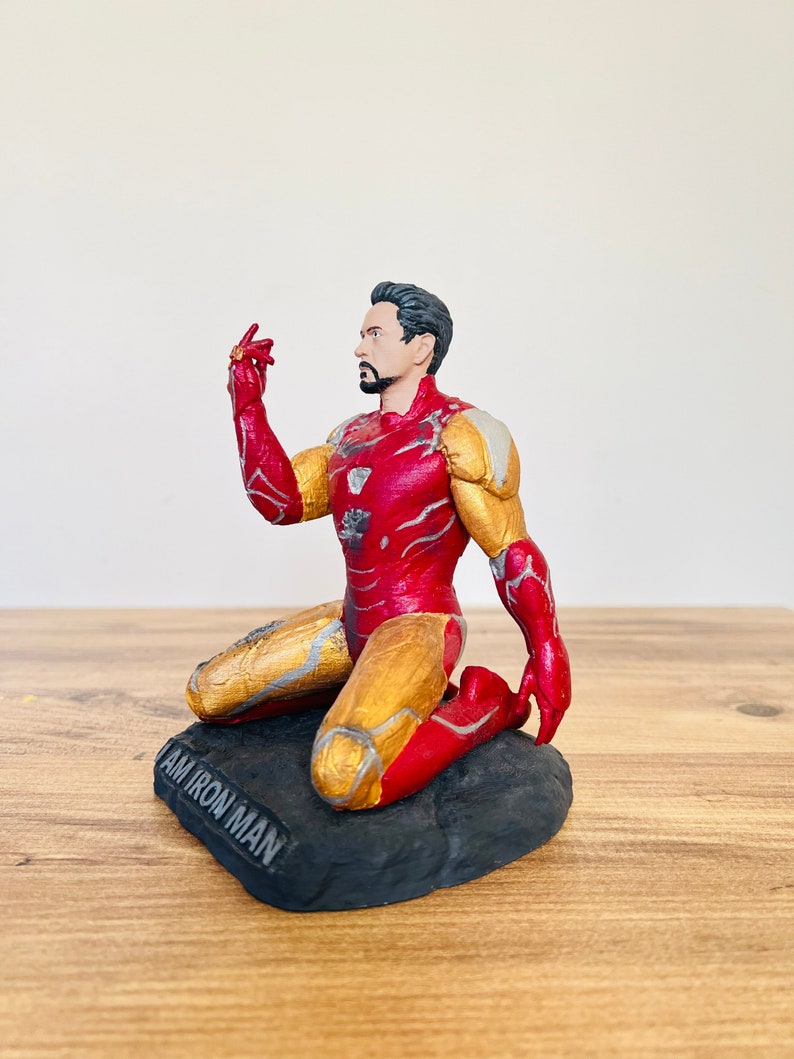 Pu&ograve; includere: Una action figure di Iron Man rossa e dorata inginocchiata su una base nera con le parole "I AM IRON MAN" incise sulla base.