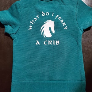 May include: A teal blue t-shirt with a white horse silhouette and the text "What do I fear? A crib" printed on the back.