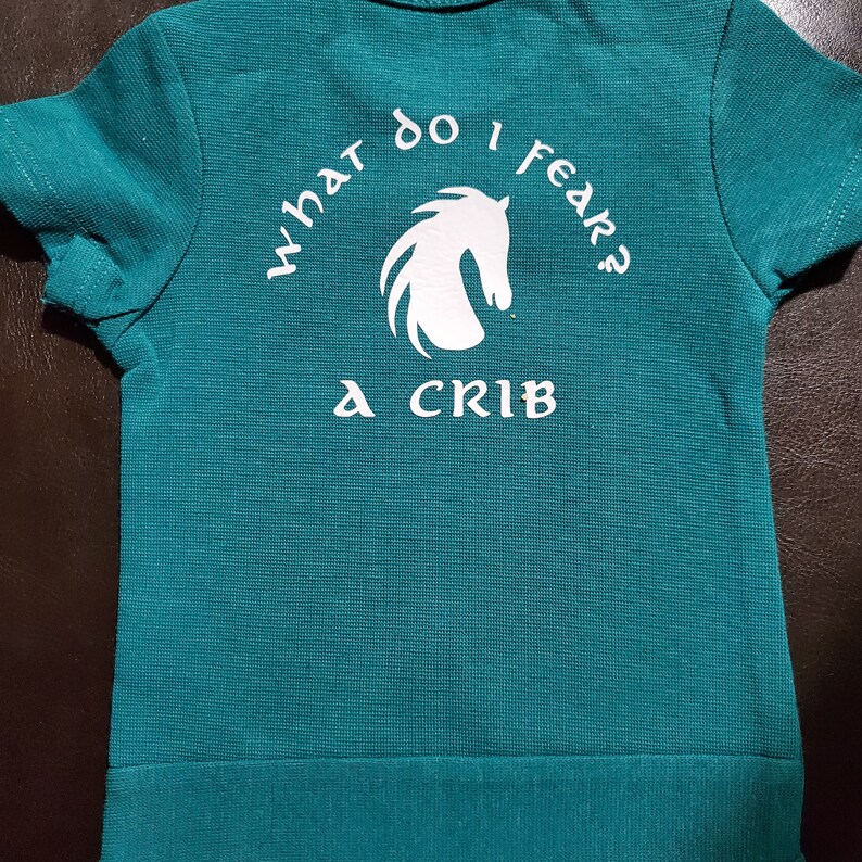 May include: A teal blue t-shirt with a white horse silhouette and the text "What do I fear? A crib" printed on the back.