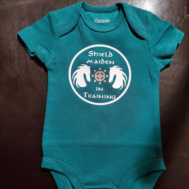 May include: Teal baby onesie with a white circle graphic that says "Shield Maiden in Training" with a stylized image of two horses' heads facing each other.