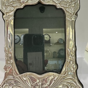 May include: A rectangular silver-toned picture frame with a scalloped edge, featuring embossed floral motifs at the top and a prominent butterfly design at the base. The frame has a polished, reflective metallic finish and a dark inner border.