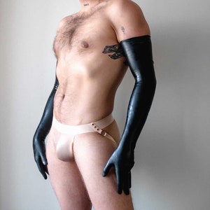 Könnte beinhalten: Schwarze Latex-Handschuhe, die bis zu den Oberarmen reichen. Ein beiger Jockstrap mit Schnallen-Detail. Das Bild zeigt eine Person mit einem Tattoo am Oberarm.