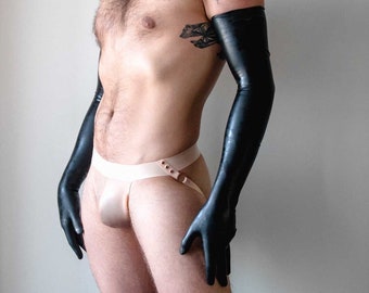 May include: Black latex gloves extending to the upper arms. A beige jockstrap with a buckle detail. The image features a person with a tattoo on their upper arm.
