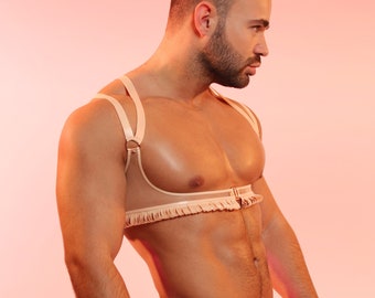 May include: A beige harness and thong set. The harness features shoulder straps, a chest band with fringe, and a connecting ring. The thong has a waistband and fringe detail. The set is made of a soft, flexible material.