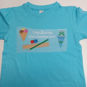May include: A turquoise t-shirt featuring a graphic with an ice cream cone, school supplies, and a school cone. The text "Hey Schule, hier komme ich!" is also printed on the shirt. The shirt is made of a soft material.