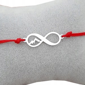 Puede incluir: Un brazalete de cuerda roja con un dije de plata en forma de símbolo de infinito que presenta una silueta de montaña.