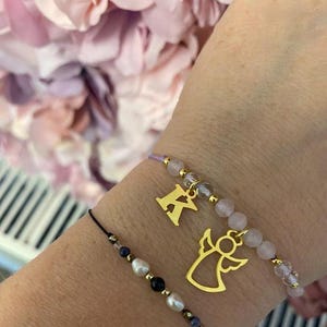 Könnte beinhalten: Zwei Armbänder mit Anhängern. Ein Armband hat einen goldenen Engelsanhänger und rosa und klare Perlen. Das andere Armband hat einen goldenen Anhänger mit dem Buchstaben K und schwarze, weiße und braune Perlen.