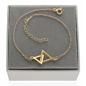 Puede incluir: Un brazalete de cadena dorada con un colgante de doble triángulo. El brazalete está en una caja blanca con un interior gris.
