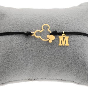 May include: A gold-colored charm bracelet featuring a black cord. The bracelet has a Mickey Mouse silhouette charm and a separate charm with the letter "M". The bracelet is displayed on a gray, plush pillow.