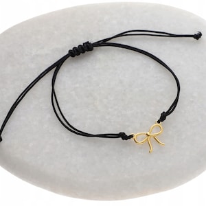 Puede incluir: Un brazalete ajustable de cordón negro con un dije de lazo dorado.