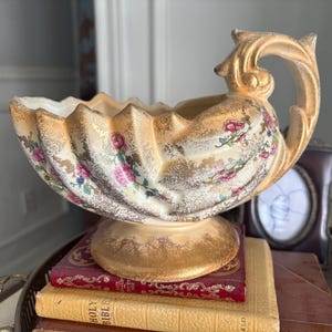 May include: A cream-colored ceramic bowl with a pedestal base and a single ornate handle. The bowl features a scalloped rim, gold-toned sponged detailing, and a floral pattern of pink roses and green leaves across the exterior surface.