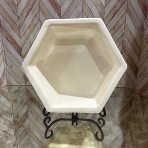 May include: A cream-coloured, hexagonal ceramic planter on a decorative black metal stand. The planter has a geometric design and a glossy finish. The stand features scrolled details, adding elegance to the piece.