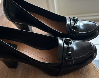 May include: A pair of black leather heeled shoes with a gold-toned buckle detail. The shoes have a pointed toe and a low block heel.