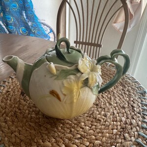 Puede incluir: Una tetera de cerámica con diseño floral. La tetera es principalmente blanca con detalles verdes y amarillos, incluyendo una flor en la parte delantera y hojas verdes. El asa y el pico también son verdes. La tetera está sobre una estera tejida.