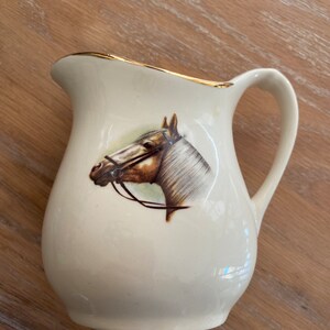 Pode incluir: Um cremoso de cer&acirc;mica branca com uma borda dourada e uma imagem pintada de uma cabe&ccedil;a de cavalo. O cavalo &eacute; marrom e branco com uma r&eacute;dea preta.