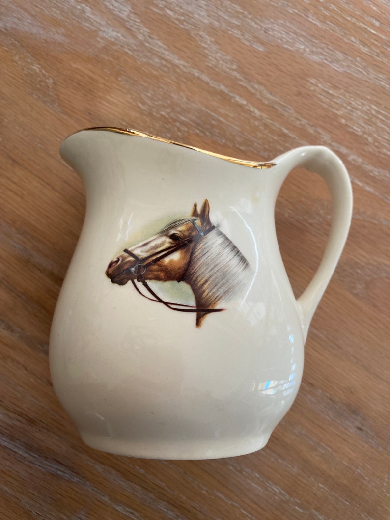 Pode incluir: Um cremoso de cer&acirc;mica branca com uma borda dourada e uma imagem pintada de uma cabe&ccedil;a de cavalo. O cavalo &eacute; marrom e branco com uma r&eacute;dea preta.