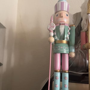 Puede incluir: Figura de cascanueces de madera en colores pastel. El cascanueces lleva una chaqueta verde claro, pantalones rosas y botas azules. Sostiene un bastón rosa con un adorno de caramelo. El sombrero es de rayas rosas y azules.