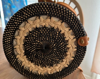 May include: A round, woven bag with a black and natural straw pattern. The bag has a brown leather strap and a gold metal clasp.