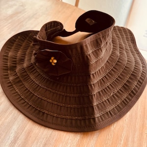 May include: A brown sun visor hat with a wide brim and a decorative flower. The hat has a structured design with horizontal stitched lines. The flower has a cluster of small, golden beads at its center. The hat is designed to provide shade from the sun.