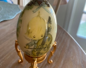 Puede incluir: Un huevo de porcelana con un soporte dorado y una imagen pintada de un ángel tocando el violonchelo. El huevo tiene una corona dorada en la parte superior.