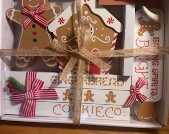 May include: A boxed gingerbread-themed gift set. It includes a gingerbread man, a gingerbread house, a rolling pin, and decorative blocks. The items are brown, white, and red, tied with twine and striped bows. The blocks read "Gingerbread Cookie Co."