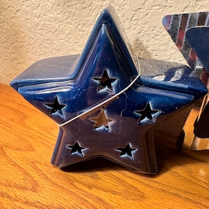 Puede incluir: Un portavelas de cerámica azul oscuro en forma de estrella con recortes de estrella. El portavelas está sobre una superficie de madera. La estrella tiene un acabado brillante y está diseñada para contener una vela en su interior.