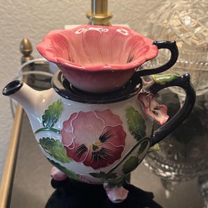 Puede incluir: Juego de té de cerámica con tetera y taza. La tetera es blanca con un diseño floral en rosa, verde y morado. La taza es rosa y se coloca encima de la tetera. El asa y el pico de la tetera son de color azul oscuro.