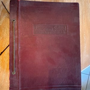 May include: A dark maroon, rectangular binder with a textured cover. The front features an embossed rectangular frame containing the text American School CHICAGO - U.S.A. A thin, vertical metal rod runs along the left side, securing the binding.