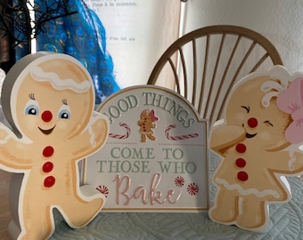 May include: Three decorative gingerbread-themed items. Two gingerbread people flank a sign that reads "Good Things Come To Those Who Bake." The gingerbread people have red buttons and smiling faces. The sign has a light pink and white color scheme.