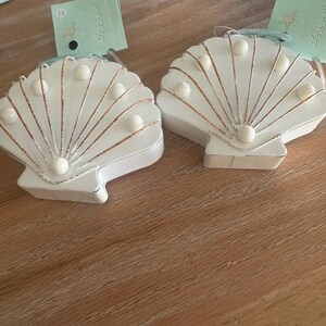 Peut inclure: Deux ornements en forme de coquillage blanc avec des accents dorés et des perles. Chaque coquillage porte une étiquette avec l'inscription "Seajelly". Les ornements sont sur une surface en bois brun clair.