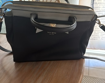 Może przedstawiać: Czarno-biała skórzana torba typu tote z złotym zamkiem błyskawicznym i złotym logo Kate Spade New York. Torba ma górny uchwyt i pasek na ramię.