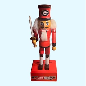 May include: A wooden nutcracker in the form of a soldier, dressed in red with a black and white hat. The hat features a large "G" logo. The nutcracker holds a sword and stands on a red base with "GEORGIA BULLDOGS" text.