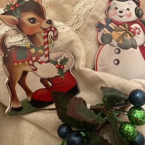 Könnte beinhalten: Vintage Weihnachtsschmuck mit einem Rentier mit Zuckerstange und einem Schneemann mit Geschenken. Das Rentier trägt einen Stechpalmenkranz und steht in der Nähe eines roten Stiefels. Ein Zweig Beeren mit blauen und grünen Akzenten ist ebenfalls vorhanden.