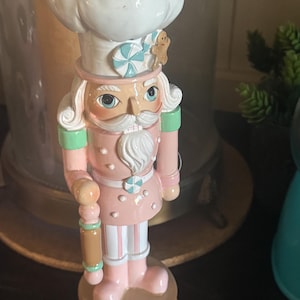 May include: A decorative nutcracker figurine in a chef's uniform. The figurine is primarily pink with white and light blue accents. It features a white chef's hat, a gingerbread man, and a candy decoration. The nutcracker is holding a rolling pin.