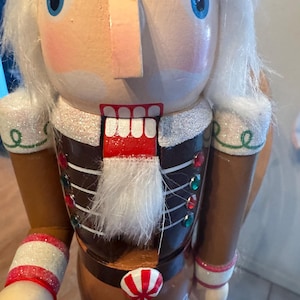 Könnte beinhalten: Ein Nussknacker aus Holz mit weißem Bart und Schnurrbart, der eine braune und weiße Uniform mit roten und weißen Zuckerstangenakzenten trägt. Der Nussknacker hat blaue Augen und einen roten Mund.