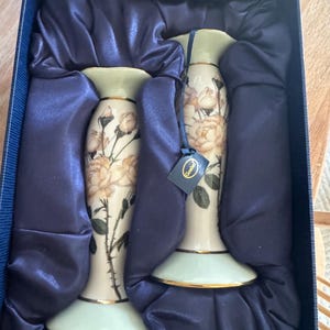 Op de afbeelding: Een paar decoratieve vazen met bloemmotieven, gepresenteerd in een blauwe geschenkdoos. De vazen hebben een crèmekleurige basis, gouden randen en een gedetailleerd rozenpatroon. De doos is gevoerd met donkerblauwe satijnstof.