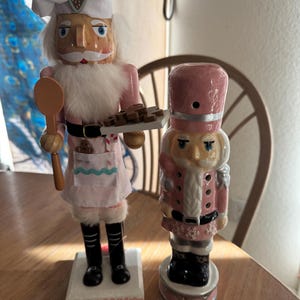 Könnte beinhalten: Zwei dekorative Nussknacker. Einer ist ein Koch mit weißer Mütze, rosa Jacke und Schürze, der einen Holzlöffel und ein Tablett mit Keksen hält. Der andere ist ein rosa-weißer Soldaten-Nussknacker mit hohem Hut und schwarzen Stiefeln.
