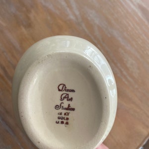 Pode incluir: Jarro de cer&acirc;mica branca com al&ccedil;a marrom e uma inscri&ccedil;&atilde;o marrom na parte inferior que diz "Dixon Art Studio 22 KT Gold U.S.A."