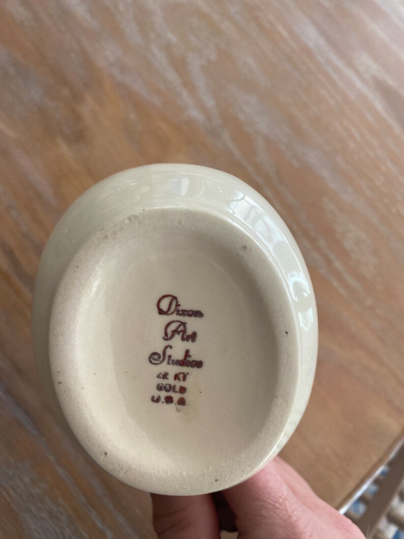 Pode incluir: Jarro de cer&acirc;mica branca com al&ccedil;a marrom e uma inscri&ccedil;&atilde;o marrom na parte inferior que diz "Dixon Art Studio 22 KT Gold U.S.A."