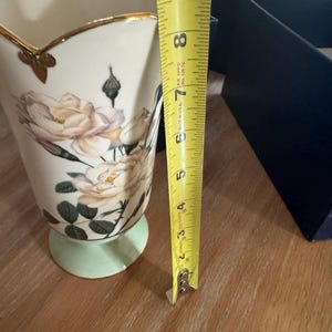 Op de afbeelding: Een crèmekleurige vaas met een bloemenrozenontwerp en gouden rand. De vaas is ongeveer 18 cm hoog en heeft een lichtgroene basis. De vaas staat op een houten oppervlak.