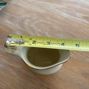 Pode incluir: Um creamer de cer&acirc;mica branca com uma borda dourada e um design floral dourado. O creamer est&aacute; sendo medido com uma fita m&eacute;trica amarela que indica "2 p&eacute;s" e "3 p&eacute;s".