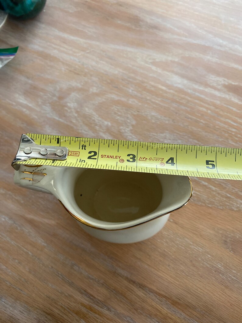 Pode incluir: Um creamer de cer&acirc;mica branca com uma borda dourada e um design floral dourado. O creamer est&aacute; sendo medido com uma fita m&eacute;trica amarela que indica "2 p&eacute;s" e "3 p&eacute;s".